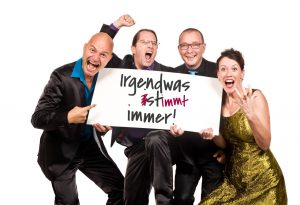 Vier Personen mit Schild 'Irgendwas stimmt immer!' in fröhlicher Stimmung.