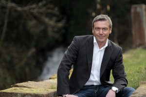 Ein Mann mit Hemd und Sakko sitzt auf einem Stein mit einem Wasserfall im Hintergrund.