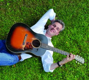 Mann liegt im Gras und hat eine Gitarre in der Hand.