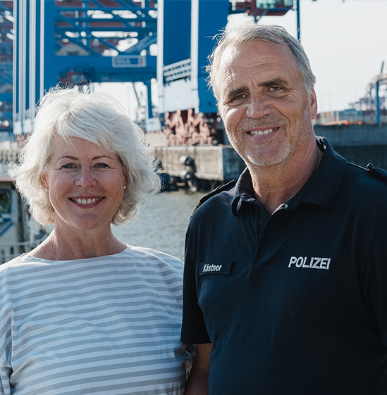Links eine Dame mit grauen halblangen Haaren, rechts danehmen ein Herr mit einer Polizei-Uniform. Im Hintergrund ist ein Hafen zu erkennen.