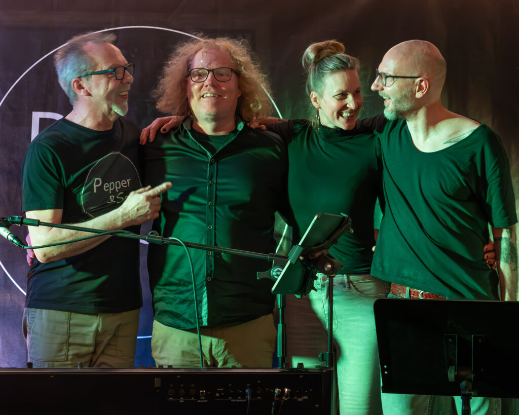 drei Männer und eine Frau mit Pferdeschwanz Arm in Arm auf einer Bühne