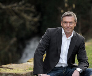 Ein Mann mit Hemd und Sakko sitzt auf einem Stein mit einem Wasserfall im Hintergrund.