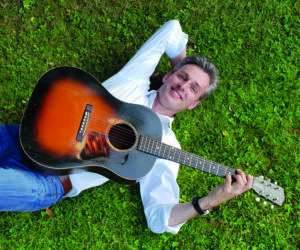 Mann liegt im Gras und hat eine Gitarre in der Hand.