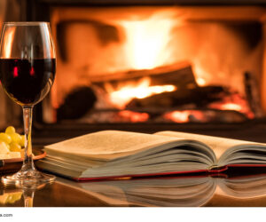 Red wine glass with an open book on table in front of burning fireplace.
