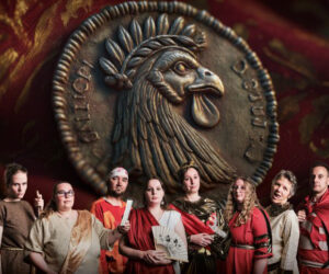 Eine Reihe von Theaterschauspieler*innen im Hintergrund eine goldene Münze mit einem Adler abgebildet