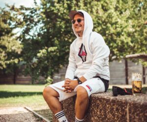 Junger Mann mit Cap und Hoodie sitz auf einer Mauer, daneben steht ein Glas Pils
