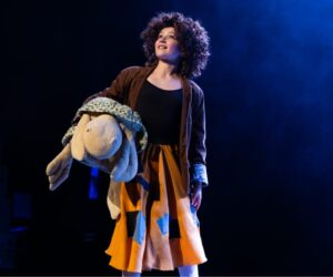 Ein Kind mit dunkelbraunen, lockigen Haaren und einem bunten Rock hält einen großen Teddy im Arm